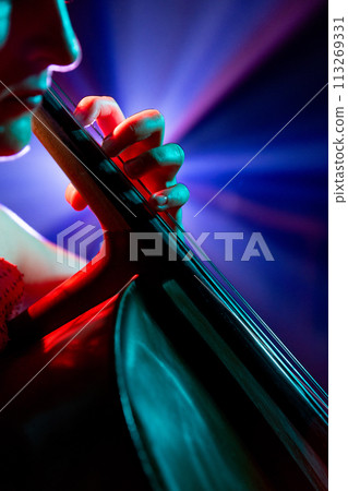 Close up, cropped portrait of musician seated, deeply focused on her cello performance under spotlights on stage. 113269331