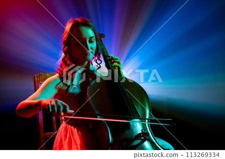 Portrait of female cellist performing classical melodies with radiant blue and purple stage lights behind her on stage. Portrait of female cellist performing classical melodies with radiant blue and purple stage lights behind her on stage. 113269334