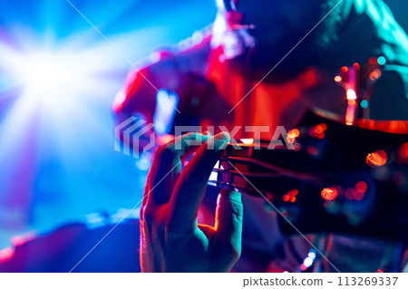 Close-up of musician's fingers on bass guitar fretboard playing melodic chords against vibrant light show. Music festivals. 113269337