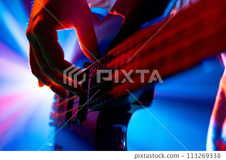 Guitarist's hands plucking strings of bass, electric guitar with dynamic stage lighting on blurred background. Guitarist's hands plucking strings of bass, electric guitar with dynamic stage lighting on blurred background. 113269338