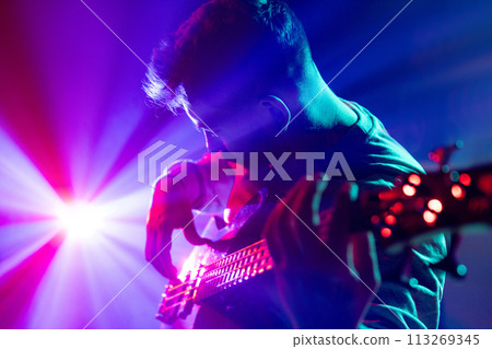 Portrait of young man performing playing chords electric guitar in neon light and visible smoke on stage. Rock and roll. 113269345
