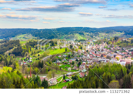 A vibrant springtime panorama over Smrzovka town, nestled amidst lush green hills and forests under a partly cloudy sky. Czechia 113271362