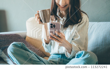 Smiling woman sitting on the sofa and using a smartphone Smiling woman sitting on the sofa and using a smartphone 113272241