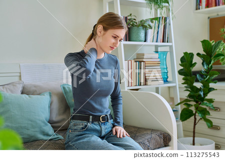 Young teenage female experiencing neck pain sitting on couch at home Young teenage female experiencing neck pain sitting on couch at home 113275543