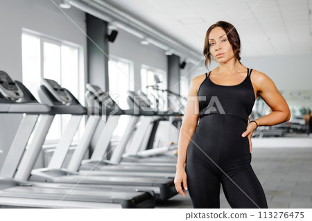 Happy beautiful fit woman is looking at camera after gym exercise. Middle aged woman wearing sport suit is standing in gym 113276475