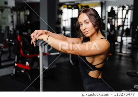 Female bodybuilder with long hair posing in gym with sports equipment. Strong woman with muscular body leaning on barbells in gym in dark atmosphere, looking at camera. Female bodybuilder with long hair posing in gym with sports equipment. Strong woman with muscular body leaning on barbells in gym in dark atmosphere, looking at camera. 113276477