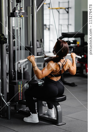 Muscular athletic female bodybuilder in black suit pulled on sports simulator in gym. Back muscles are very tense. Muscular athletic female bodybuilder in black suit pulled on sports simulator in gym. Back muscles are very tense. 113276478