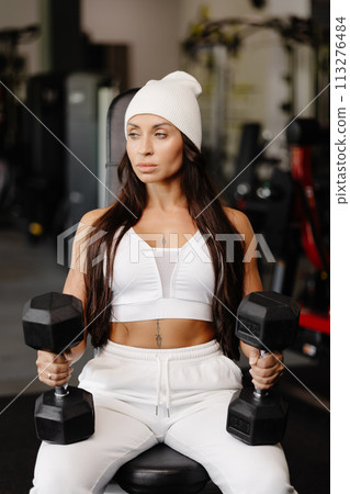 Athletic young fitness woman is wearing in white sport suit in gym outfit doing shoulder workout with dumbbells 113276484