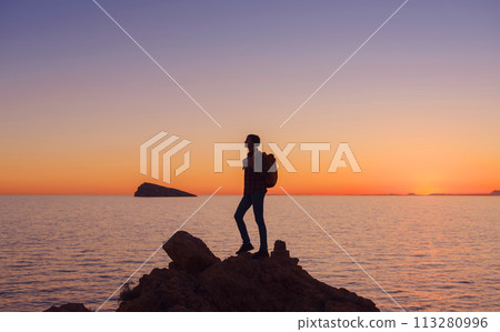 silhouette of Young woman hiking on rocky beach in Spain, Benidorm. Watching the choppy sea and the bay. traveler enjoying freedom in serene sunset landscape, success concept 113280996