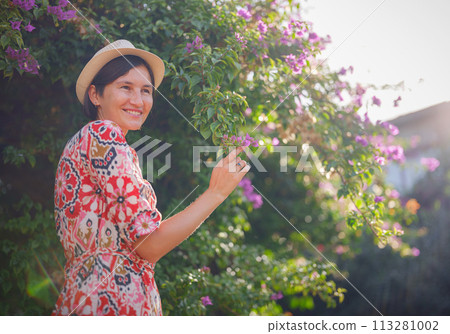 young asian woman stands in sunny green park, admiring blooming bougainvillea bush. The flower's bright colors are beautiful contrast to park's lush greenery. young asian woman stands in sunny green park, admiring blooming bougainvillea bush. The flower's bright colors are beautiful contrast to park's lush greenery. 113281002