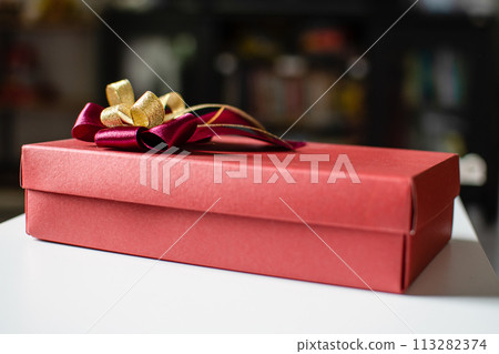 The red color Presents celebrate box with red and gold ribbons placed on a white table with copy space on the top can input text, take close-up photography, and top view, blurred background 113282374