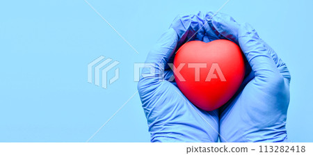 Both hands were wearing gloves and holding a red heart. Concept world heart foundation organizes world heart day, copy space on left for design or text, close-up, blurred blue background Both hands were wearing gloves and holding a red heart. Concept world heart foundation organizes world heart day, copy space on left for design or text, close-up, blurred blue background 113282418