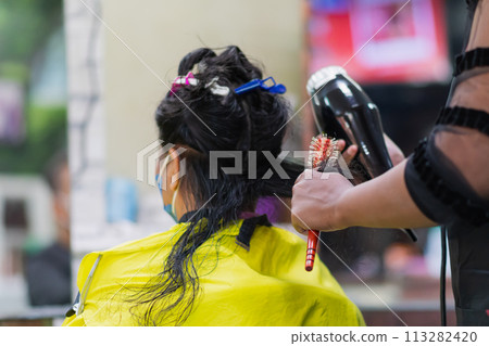 professional woman hairdresser wearing face mask holding hair dryer and hair styling client woman indoor salon, Business hair care has tool haircut to service haircut to a customer, selective focus professional woman hairdresser wearing face mask holding hair dryer and hair styling client woman indoor salon, Business hair care has tool haircut to service haircut to a customer, selective focus 113282420