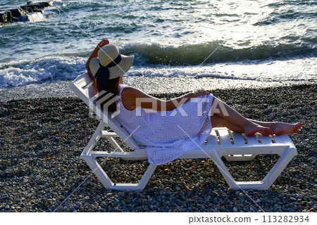 a girl in a hat and a summer white dress lies on the beach and looks into the distance a girl in a hat and a summer white dress lies on the beach and looks into the distance 113282934
