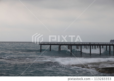 A lone figure in front of foam waves and a historic mountain in Alanya, November 2021. 113283353