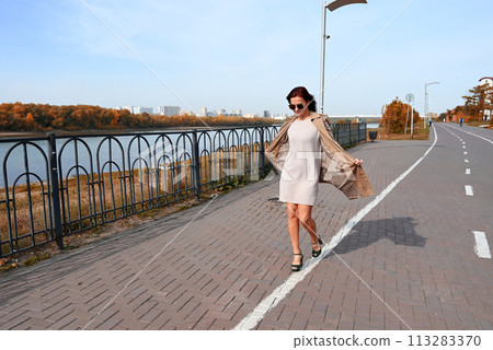a confident young attractive woman in a coat and high-heeled shoes walks through the streets of the city, the woman smiles and enjoys life 113283370