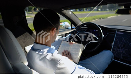 Male businessperson reading book during riding on electrical vehicle with autopilot at urban road. Successful businessman improving his knowledge while riding an autonomous self driving electric car 113284353