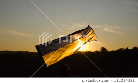 Young man in military uniform waving flag of Ukraine against beautiful sunset at background. Male ukrainian army soldier lifted national banner at countryside. Victory against russian aggression 113284381