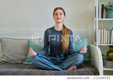 Young teenage relaxed female sitting in lotus position, meditating at home 113284755