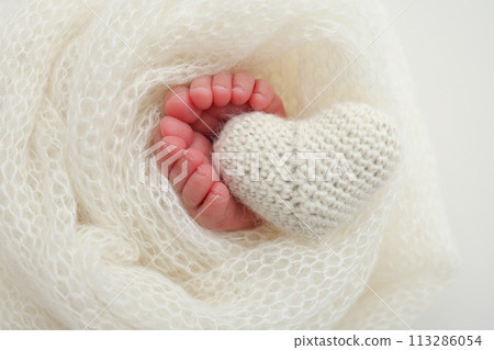 The tiny foot of a newborn baby. Soft feet of a new born in a white wool blanket. Close up of toes, heels and feet of a newborn. Knitted white heart in the legs of a baby. Macro photography 113286054