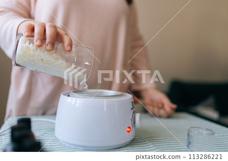 Close-up hands of unrecognizable female artisan craftperson pouring dry ingredient from glass measuring jar for creating candle building mixture. Process of making handmade natural scented candle. Close-up hands of unrecognizable female artisan craftperson pouring dry ingredient from glass measuring jar for creating candle building mixture. Process of making handmade natural scented candle. 113286221