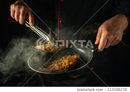 The chef is frying fish in the kitchen. The concept of preparing a fish dish for lunch. The cook hand takes out hot fried crucian carp from the frying pan with tongs 113286230