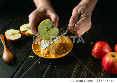 Chef hands with a grater grate an apple on the kitchen table. Low key concept of preparing a healthy fruit dinner 113286232