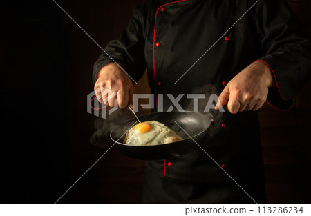 Cooking scrambled eggs in a frying pan in the hands of a chef. Low key concept of preparing a delicious breakfast with chicken eggs. Grande cuisine Cooking scrambled eggs in a frying pan in the hands of a chef. Low key concept of preparing a delicious breakfast with chicken eggs. Grande cuisine 113286234