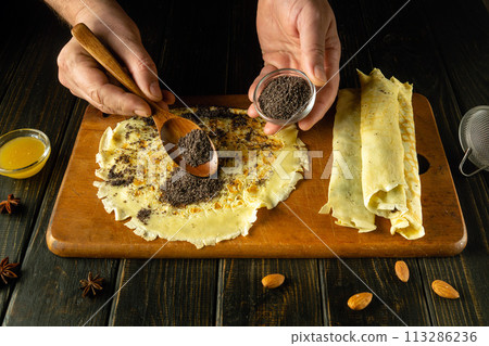 Close-up of a man hands adding poppy seeds to a pancake with a spoon. Preparing a sweet dessert with poppy seeds and honey on the kitchen table 113286236