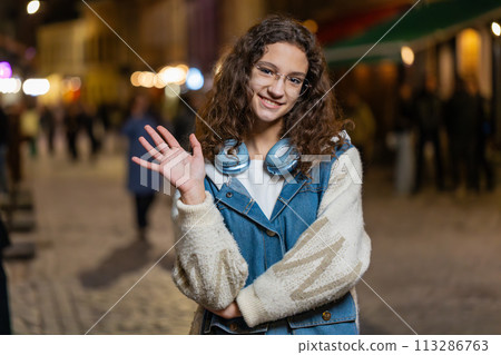 Teenage girl waving hands gesturing hi hello greeting or goodbye on urban city street at night Teenage girl waving hands gesturing hi hello greeting or goodbye on urban city street at night 113286763
