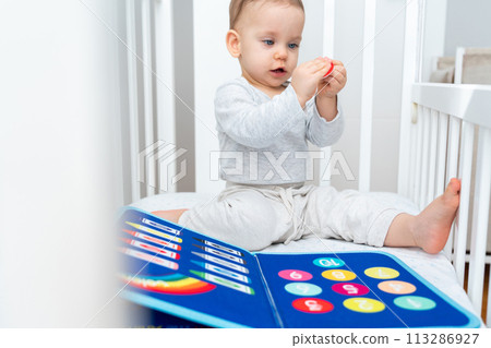 Baby sitting in a crib playing with montessori busy book. Concept of keeping children from screen by activity books and quiet books Baby sitting in a crib playing with montessori busy book. Concept of keeping children from screen by activity books and quiet books 113286927