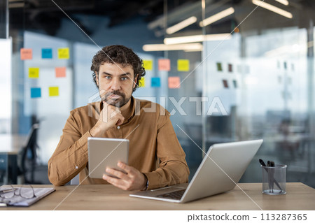 Focused professional using a laptop and a tablet at a wooden desk in a modern office, with sticky notes in the background. 113287365
