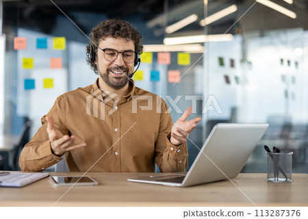A happy bearded man wearing headphones talks cheerfully in a video conference in a modern office setting. 113287376