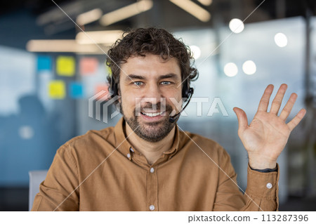 Portrait of a cheerful male customer service representative wearing a headset and waving in a modern office setting Portrait of a cheerful male customer service representative wearing a headset and waving in a modern office setting 113287396