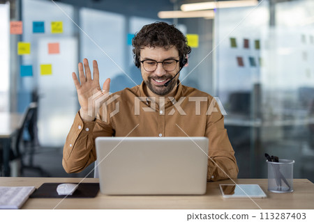 Professional man in a casual business setting using laptop for a cheerful video conference with colleagues or clients, waving to the camera. Professional man in a casual business setting using laptop for a cheerful video conference with colleagues or clients, waving to the camera. 113287403