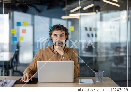 Focused Hispanic male entrepreneur using laptop in a contemporary workspace, looking at the camera. 113287409