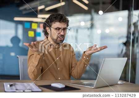 An adult male in a casual shirt expresses confusion while using a laptop in a contemporary office setting, reflecting common workplace challenges. 113287429