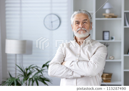 A portrait of a confident senior man with folded arms standing in a stylishly furnished home interior showing contentment and maturity. A portrait of a confident senior man with folded arms standing in a stylishly furnished home interior showing contentment and maturity. 113287490