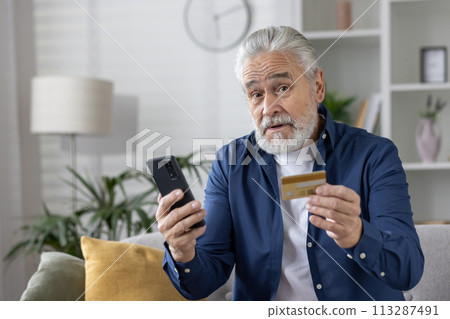 Elderly male with credit card and phone, potential victim of online fraud in a cozy home setting. 113287491