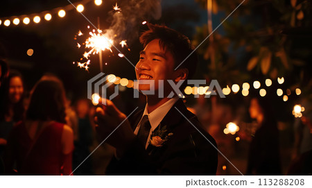 A cheerful young Asian guy in a formal suit holds a sparkler at an outdoor evening party, illuminating the faces in the crowd with a warm glow. Formal event, school graduation A cheerful young Asian guy in a formal suit holds a sparkler at an outdoor evening party, illuminating the faces in the crowd with a warm glow. Formal event, school graduation 113288208