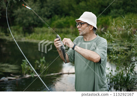 Man with fishing rod near the lake fishing at summer. Fisherman catch a fish. Man wearing khaki clothes and a cap. 113292167