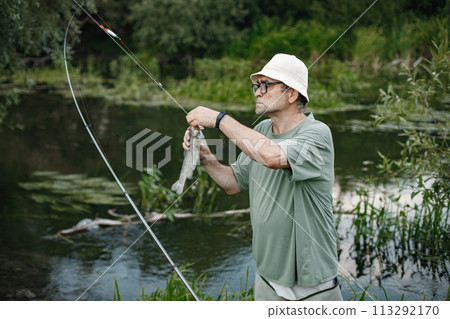 Man with fishing rod near the lake fishing at summer. Fisherman catch a fish. Man wearing khaki clothes and a cap. 113292170