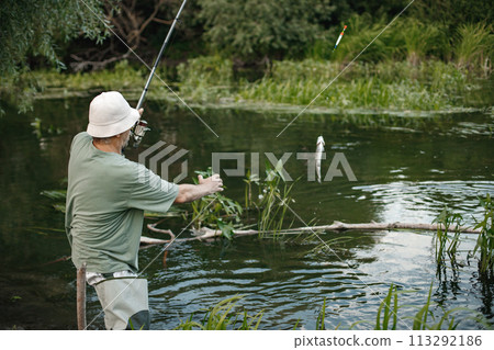 Man with fishing rod near the lake fishing at summer. Fisherman catch a fish. Man wearing khaki clothes and a cap. Man with fishing rod near the lake fishing at summer. Fisherman catch a fish. Man wearing khaki clothes and a cap. 113292186
