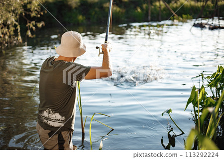Man with fishing rod near the lake fishing at summer. Fisherman enjoying his hobby. Man wearing khaki clothes and a cap. 113292224