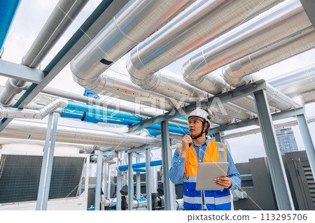 engineer man in a safety vest is standing around gas plump pipe, looking at a laptop. He is wearing a hard hat and a helmet engineer man in a safety vest is standing around gas plump pipe, looking at a laptop. He is wearing a hard hat and a helmet 113295706