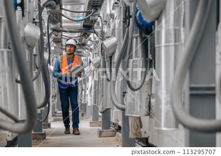 man in a safety vest is walking through a large industrial building with pipes and wires. He is holding a clipboard inspecting pipeline area. man in a safety vest is walking through a large industrial building with pipes and wires. He is holding a clipboard inspecting pipeline area. 113295707