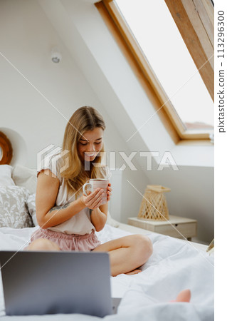 Young woman is sitting on a bed with a laptop and a cup of coffee 113296300