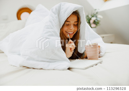 A woman lies on the bed under a blanket, smiling as she writes in her notebook 113296328