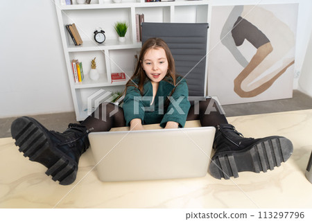 Little cheerful girl sitting with laptop with her feet on the table. High quality photo 113297796