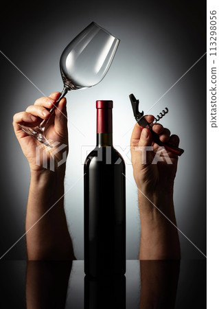 Unopened bottle of red wine and hands with corkscrew and wine glass. 113298056